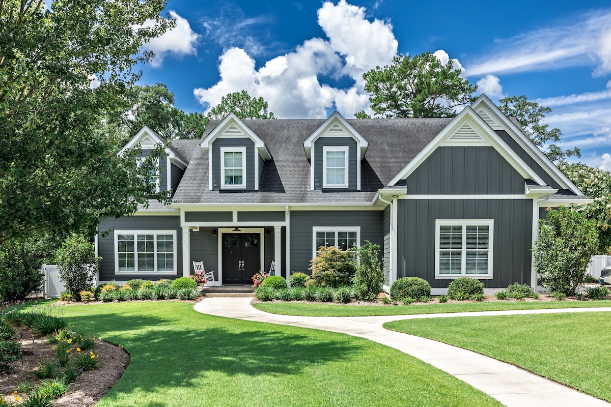 A large gray craftsman new construction house with a landscaped yard and leading pathway sidewalk on a sunny day with blue skies and clouds.