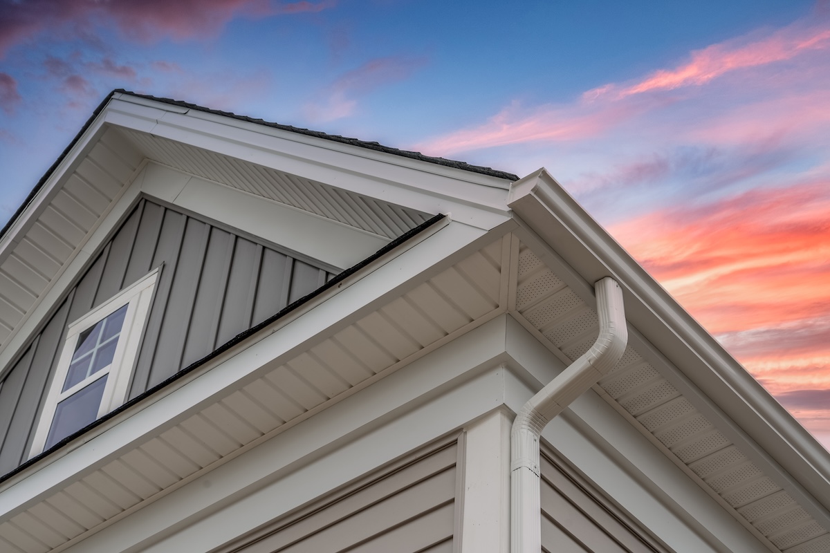 White frame gutter guard system, with gray horizontal and vertical vinyl siding fascia, drip edge, soffit, on a pitched roof attic at a luxury American single family home dramatic sunset sky