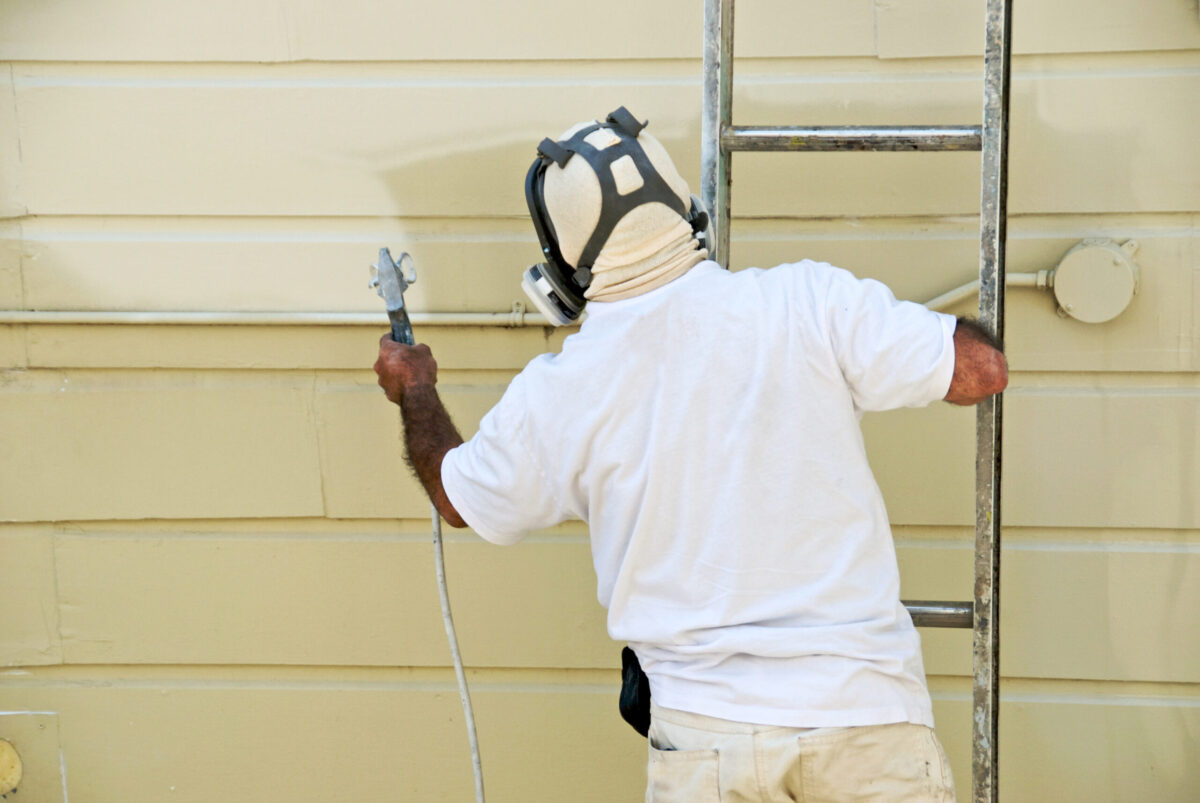 A man on a ladder uses spray gun to paint the exterior of a wooden building how to paint vinyl siding
