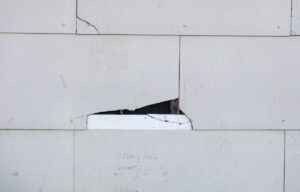 Asbestos siding falling apart due to age. Main focus is on major damage shingle with felt paper underneath.