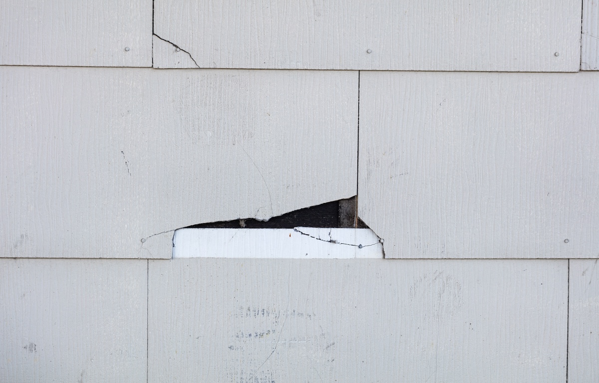 Asbestos siding falling apart due to age. Main focus is on major damage shingle with felt paper underneath.