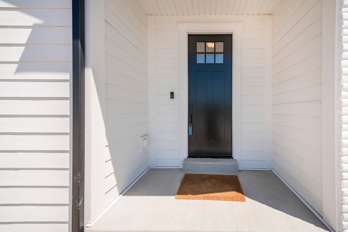 Exterior of a house with a view of black wooden door with glass panel and an enginered wood sidings. There is a doorbell beside the door and a brown doormat on the floor. engineered wood siding
