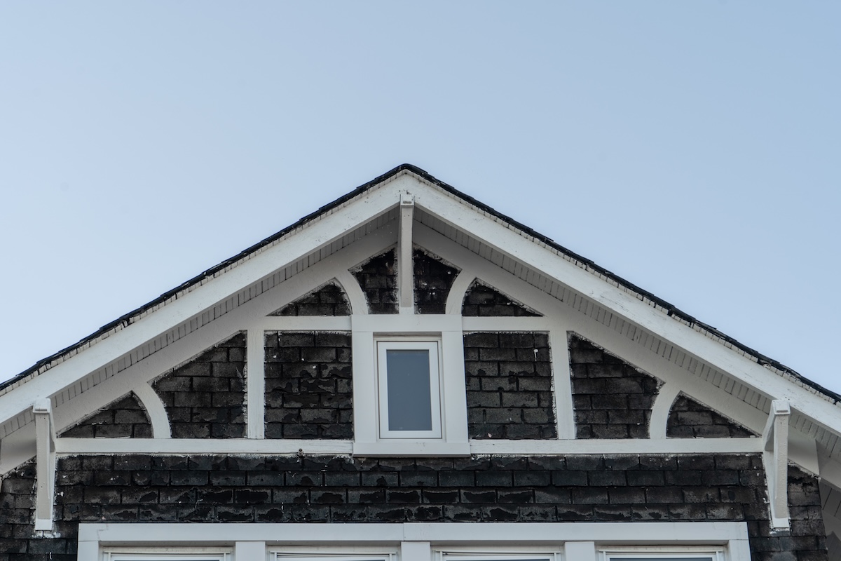 House facade covered in dark shake and shingle white, dividers, gable held by decorative brackets, white attic square window black siding