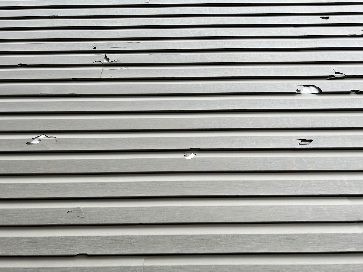 Holes in the exterior siding of the home from hail storm damage. The broken vinyl siding on a house due to hail.